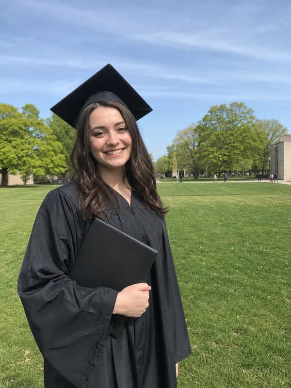 Photo of a graduate in cap and gown on a campus lawn holding her diploma